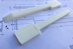 Frezowane prototypy z tworzywa sztucznego (POM:PEEK) leżące na rysunku technicznym. Produkcja narzędzi i przyrządów.